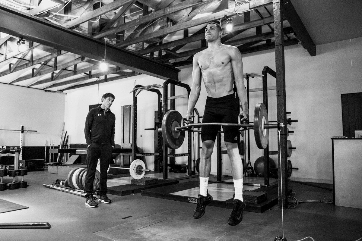 Athlete at the peak of a loaded trap bar jump with a P3 coach observing, black and white